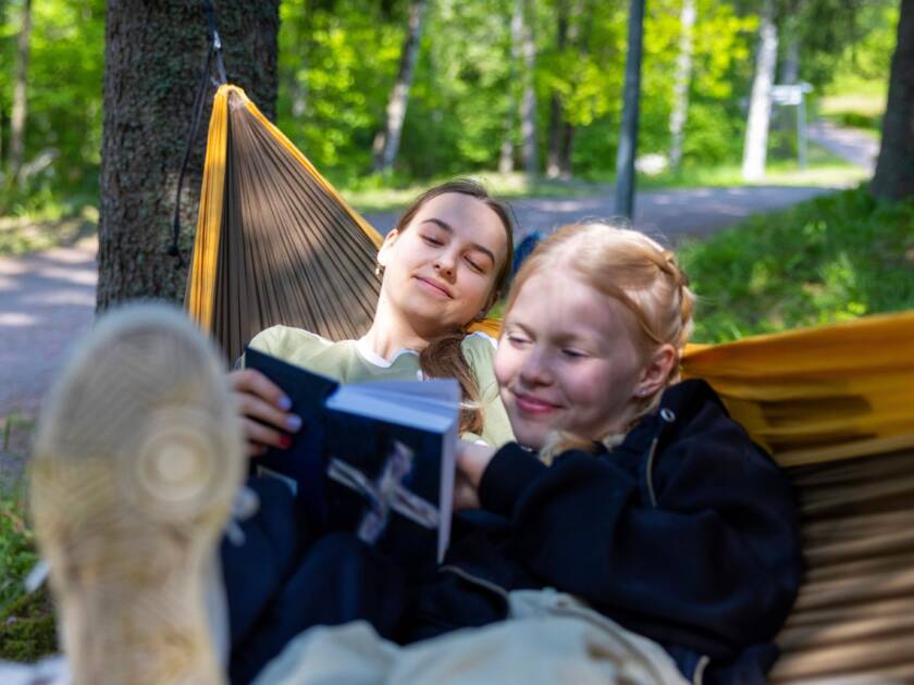 Kaksi nuorta naista makaa riippumatossa metsässä, lukien Raamattua ja hymyillen yhdessä. Taustalla vihreitä puita ja polku.