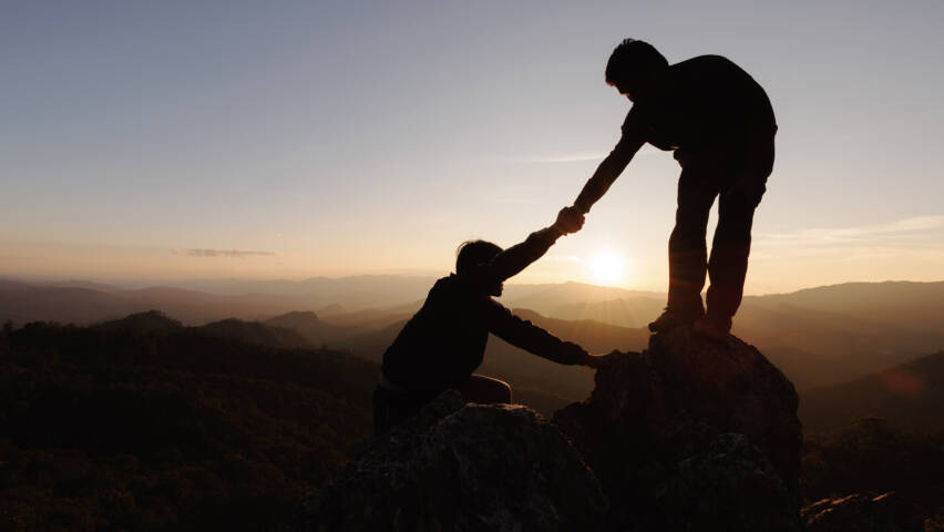 Silhouettes of two people climbing on mountain and helping each other.